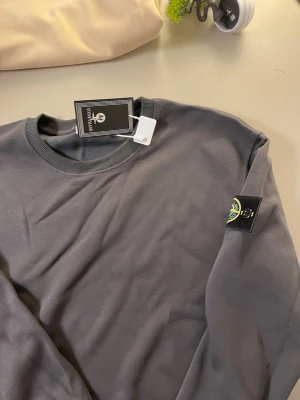 Mörkgrå sweatshirt från Stone Island - Snygg mörkgrå sweatshirt från Stone Island med klassisk rund halsringning och ribbade muddar. Tröjan har den ikoniska Stone Island-loggan på vänster ärm och är tillverkad i mjukt bomullsmaterial. Perfekt för en avslappnad och trendig stil.