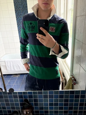 Randig rugbytröja från Morris Gents - Snygg långärmad rugbytröja från Morris Gents med breda gröna och marinblå ränder, vit krage och patchdetaljer på bröstet. Tröjan har knappar i halsen och förstärkta armbågar i brunt. Perfekt för en sportig och avslappnad stil.