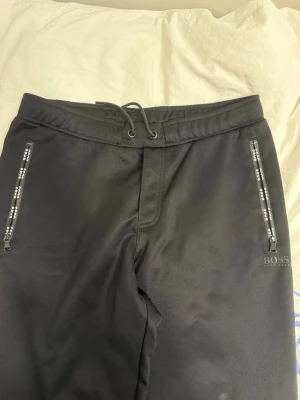Svarta mjukisbyxor från BOSS - Svarta mjukisbyxor från BOSS med dragsko i midjan och dragkedjor på fickorna där det står BOSS. Byxorna har en stilren look och är perfekta för en avslappnad stil. Materialet känns mjukt och bekvämt.