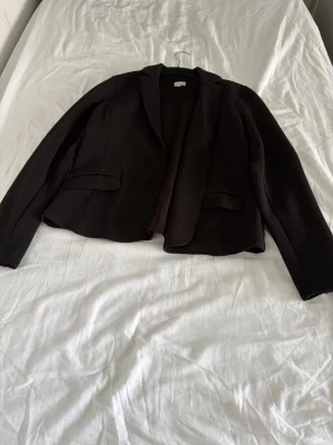 Svart kavaj med fickor - Stilren svart kavaj med klassisk krage och två fickor framtill. Kavajen har en rak passform och är tillverkad i ett strukturerat tyg som ger en modern känsla. Perfekt att matcha med skjorta eller t-shirt för en snygg look.