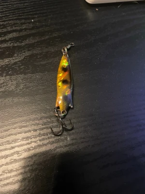 Färgglatt fiskedrag med trekrok - Säljer ett fiskedrag i metall med holografisk yta i guld, grönt och rött. Draget har en böjd form och är utrustat med en trekrok i metall. Perfekt för fiskeentusiasten som vill ha något som syns i vattnet.