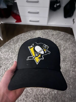 Svart Pittsburgh Penguins keps - Svart keps med Pittsburgh Penguins-logga framtill och Winter Classic-märke på sidan. Böjd skärm och broderade detaljer i gult, vitt och svart. Perfekt för hockeyfans som vill visa sitt stöd för laget.