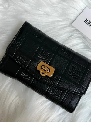 Svart plånbok från Steve Madden - Snygg svart plånbok i präglat konstläder från Steve Madden med guldigt spänne. Plånboken har flera kortfack, myntfack med dragkedja och ett fönster för ID-kort. Perfekt för att hålla ordning på dina kort och kontanter.