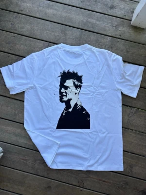 Vit Tyler durden Fight Club t-shirt med tryck - Säljer en vit t-shirt med svart tryck av Fight Club-loggan fram och en stor grafisk bild på ryggen. T-shirten har klassisk passform och korta ärmar. Perfekt för dig som gillar film och vill ha en unik look.