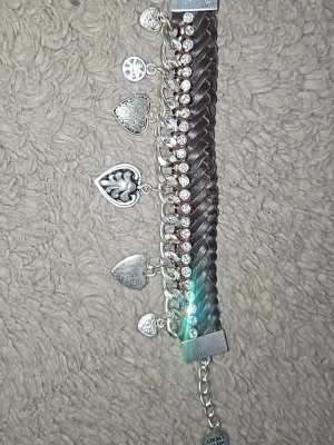 Brett armband med hjärtberlocker - Snyggt brett armband i mörkbrunt med flätad design och silverfärgade detaljer. Armbandet har flera hjärtformade berlocker och gnistrande stenar längs ena sidan. Perfekt accessoar för att lyfta din stil med en unik touch.
