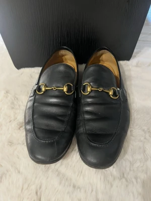 Gucci Svarta loafers  - Stilrena äkta Gucci svarta loafers i läder med klassisk guldfärgad metallspänne över vristen. Skorna har en tidlös design med rundad tå och låg klack. Insidan är fodrad i beige och de passar perfekt till både jeans och kostymbyxor.                     (Priset går att diskutera om)