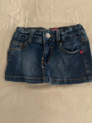 Blå jeanskjol med fickor och knapp - Snygg blå jeanskjol i kort modell med klassiska fickor fram och bak, bälteshällor och knapp framtill. Kjolen har kontrastsömmar och en liten rosa detalj vid fickan. Perfekt för dig som gillar denim och vill ha en enkel men trendig look.