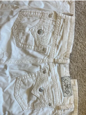Vita bootcut missme jeans - Säljer ett par vita bootcut jeans med snygga fickdetaljer och silverfärgade knappar. Jeansen har dekorativa sömmar och en klassisk femficksmodell. Materialet är troligtvis bomull med stretch för skön passform. Perfekta för dig som gillar en clean och fräsch look. jag är 170 ish och brukar ha 38cm i midjan, skulle säga att de är lite större men jag har sytt in som så att dom passar typ s/m