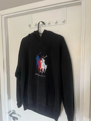 Svart hoodie med polospel-tryck - Säljer en stilren Ralph Lauren hoodie med stor huva och ett färgstarkt tryck av tre polospelare i blått, rött och vitt på bröstet. Hoodien har en klassisk känguruficka framtill och ribbade muddar vid ärmar och nederkant. Perfekt för en avslappnad och sportig stil. Storlek M passar 184-187 isch