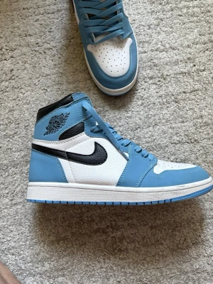 Blå och vita Air Jordan 1 High - Säljer ett par Air Jordan 1 High sneakers i blått och vitt med svarta detaljer och klassisk Nike swoosh. Skorna har perforerad tåbox, högt skaft och är gjorda i läder. Perfekta för dig som vill sticka ut med en ikonisk och stilren sneaker.