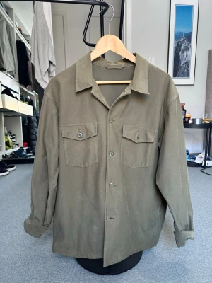 Olivgrön overshirt med bröstfickor - Stilren olivgrön overshirt med två stora bröstfickor med lock och knapp. Klassisk krage och knäppning framtill. Tillverkad i ett mjukt, lite tjockare tyg som ger en avslappnad look. Perfekt att bära öppen över en t-shirt.