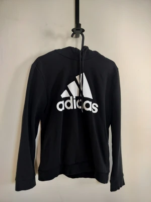 Svart hoodie med Adidas-logga - Svart hoodie från Adidas med stor vit logga och text på bröstet. Klassisk modell med huva och dragsnören, samt långa ärmar. Tillverkad i mjukt material som känns skönt mot huden. Perfekt för en avslappnad och sportig stil.
