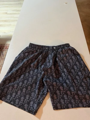 Mörkgrå mönstrade shorts med snörning - Säljer ett par mörkgrå shorts med svart grafiskt mönster och elastisk midja med snörning. Shortsen har en avslappnad passform och är perfekta för sommaren. Materialet känns lätt och bekvämt, och de har fickor både fram och bak.