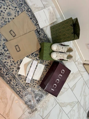 Vita Gucci sneakers med broderad detalj - Säljer ett par vita sneakers från Gucci med klassisk låg profil och snörning. Skorna har en broderad röd och grön detalj på sidan samt inslag av skinn. Kommer med originalkartong, dustbags och tillbehör.