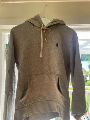 Grå hoodie från Polo Ralph Lauren - Snygg grå hoodie från Polo Ralph Lauren med klassisk huva och vita dragsnören. Har en stor magficka och den ikoniska blåa loggan broderad på bröstet. Perfekt för chill dagar och enkel att matcha med allt. Tillverkad i mjukt bomullsmaterial.