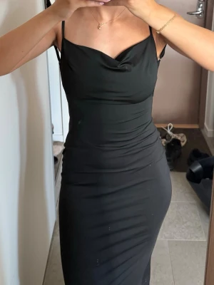 Svart långklänning med öppen rygg - Säljer en svart långklänning med smala axelband och snygg draperad urringning. Klänningen har en figurnära passform och öppen rygg som ger en elegant look. Perfekt för dig som vill ha en stilren och tidlös outfit.