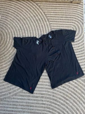 Svart t-shirt med röd logga Från Ralph Lauren  - Två svarta t-shirts från Ralph Lauren med rund hals och korta ärmar. Diskret röd broderad logga nertill på framsidan. Klassisk och enkel design som passar till det mesta. Tillverkad i mjuk bomull för skön känsla hela dagen.