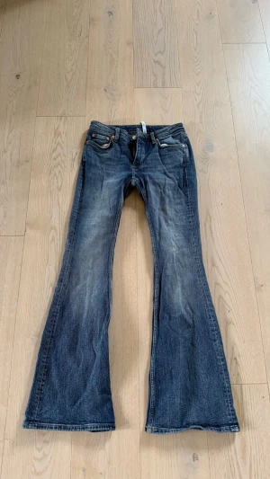 Blå bootcut jeans med slitningar - Säljer ett par blå bootcut jeans med snygga slitningar och klassisk femficksdesign. Jeansen har låg midja och vida ben, vilket ger en retro och avslappnad look. Tillverkade i denim med en tvättad finish för extra karaktär.