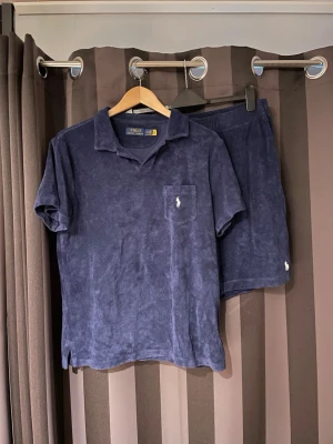 Mörkblå frottéset från Polo Ralph Lauren - Set i mörkblå frotté från Polo Ralph Lauren med kortärmad skjorta och matchande shorts. Båda delarna har broderad logga och en mjuk, avslappnad känsla. Perfekt för sommaren.