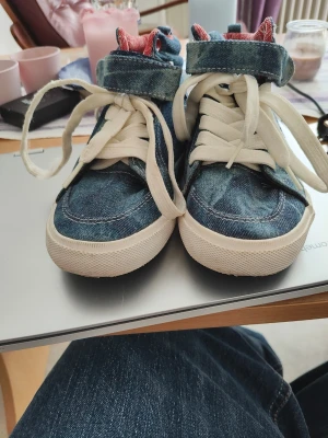 Blå denim high top sneakers - Säljer ett par unika high top sneakers i blått denimtyg med vita sulor och vita skosnören. Skorna har röda detaljer på insidan och metallöljetter. Perfekta för dig som vill sticka ut med en cool streetstyle-look.