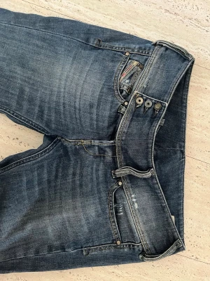 Diesel-Mörkblå bootcut jeans med låg midja - Säljer ett par mörkblå jeans med låg midja och bootcut passform. Jeansen har slitningar och tvättade detaljer framtill, samt tre knappar i gylfen. Klassisk femficksmodell i denim med kontrastsömmar.