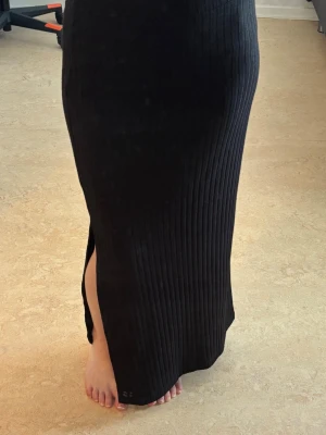 Svart ribbad långkjol med slits - Säljer en svart långkjol med ribbad struktur och slits på sidan. Kjolen har en tight passform och är perfekt för dig som gillar en stilren och enkel look. Materialet känns mjukt och följsamt mot kroppen.