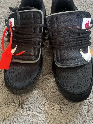 Svarta Nike Off-White sneakers - Säljer ett par svarta Nike Off-White sneakers med vit swoosh, orange detalj och röd zip-tie. Skorna har meshmaterial, tjock plös och texten 'AIR' på sidan. Snygg design med svarta skosnören och kontrasterande sömmar.
