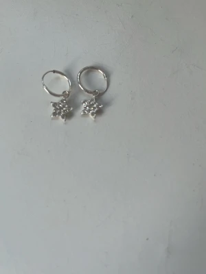  Äkta silver örhängen  - Säljer ett par silvriga örhängen med små stjärnformade berlocker. Stjärnorna är dekorerade med gnistrande stenar som ger ett elegant och trendigt intryck. Perfekta för dig som gillar detaljer som sticker ut och vill addera lite extra bling till din stil.