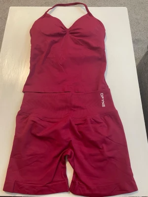 Vinrött träningsset från DFYNE - Säljer ett vinrött träningsset från DFYNE med ett linne med smala axelband och matchande shorts med hög midja. Setet är gjort i stretchigt material som passar perfekt för träning. Både linnet och shortsen har DFYNE-logga framtill.