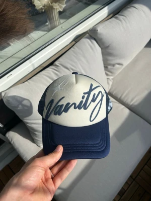 Mörkblå Trucker Keps av VanityWrld - Mörkblå och kräm vit keps från vanitywrld, en puffprint logga på framsidan med en embroidery text på sidan
