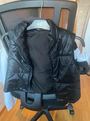 Svart dunväst med hög krage - Snygg svart dunväst från H&M med hög krage och dragkedja framtill. Västen har en klassisk puffer-look och är perfekt att slänga över en hoodie eller tröja. Materialet är glansigt och quiltat för extra stilpoäng.