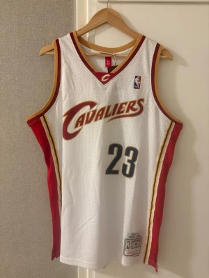 Cleveland Cavaliers basketlinne #23 - Snyggt vitt basketlinne med röda och gula detaljer från Cleveland Cavaliers. Nummer 23 och namnet James på ryggen. Luftigt meshmaterial perfekt för basket eller streetwear. NBA-logga på bröstet och officiell märkning nertill.