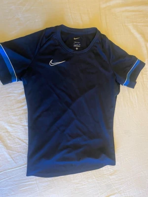 Mörkblå Nike sporttröja - Säljer en mörkblå sporttröja från Nike med korta ärmar och vita samt blå detaljer på ärmsluten. Tröjan har en vit Nike-logga på bröstet och är tillverkad i ett snabbtorkande funktionsmaterial, perfekt för träning eller sportaktiviteter.