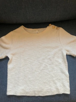Vit topp med guldknappar på axeln - Säljer en vit topp i bouclé-liknande material med rund hals och korta ärmar. På ena axeln sitter tre dekorativa guldknappar som ger en lyxig touch. Perfekt till jeans eller kjol för en trendig look.