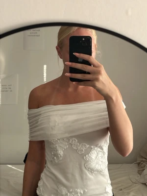 Vit offshoulder topp med spetsdetaljer - Säljer en vit offshoulder topp med bred, vikt kant över axlarna och fina broderade spetsdetaljer framtill. Toppen är figurnära och har en lätt transparent baksida. Perfekt för dig som vill ha något unikt och stilrent.