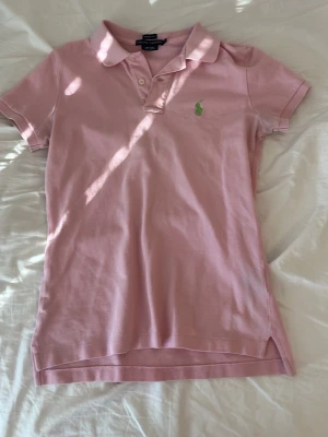 Rosa pikétröja från Ralph Lauren - Snygg rosa pikétröja från Ralph Lauren i bomull. Klassisk krage, korta ärmar och grön broderad logga på bröstet. Skinny fit-modell som sitter tajt och har knappar vid halsen. Perfekt för en fräsch och sportig look.