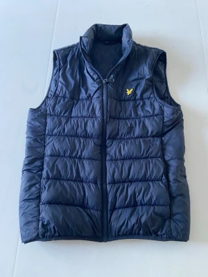 Mörkblå dunväst med gul logga - Snygg mörkblå dunväst med gul broderad logga på bröstet. Västen har hög krage, dragkedja framtill och quiltad design. Perfekt för lager-på-lager och håller dig varm under kyliga dagar.