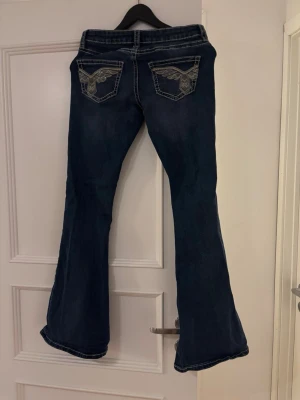 Bootcut jeans med broderade fickor - Mörkblå bootcut jeans med snygga broderade detaljer på bakfickorna och kontrastsömmar. Jeansen har låg midja och klassisk femficksdesign. Perfekta för dig som gillar en retrostil och vill ha ett par jeans som sticker ut.
