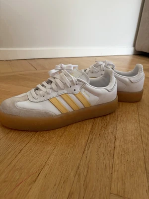 Adidas sneakers med gula ränder - Säljer ett par vita Adidas sneakers med klassiska gula ränder på sidorna och beige detaljer vid tån. Skorna har en tjock gummisula i beige. Skorna är endast använda en gång.