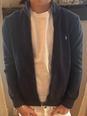 Stickad Ralf Lauren kofta  - Mörkblå stickad kofta med dragkedja framtill och ribbade muddar. Två fickor på framsidan och enkel, stilren design. Perfekt för dig som gillar en avslappnad och modern look.