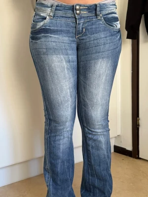Blå bootcut jeans med dubbla knappar - Säljer ett par blå bootcut jeans med dubbla knappar i midjan. Jeansen har klassiska fem fickor och ljusare tvättade partier framtill för en cool look. Perfekta för dig som gillar en avslappnad men trendig stil. Jeansen är väl använda där av slitningen vid foten. midjemått - 37 och innerbenslängd - 86