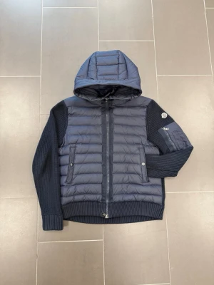 Moncler mörkblå puffer cardiganjacka - Mörkblå puffer cardiganjacka från Moncler med vadderad front och stickade ärmar. Jackan har huva, två sidofickor med knapp, ärmficka med dragkedja och Moncler-logga på vänster ärm. Materialmix av nylon och ull ger både värme och stil.