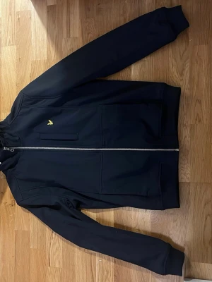 Mörkblå jacka från Lyle & Scott - En knappt använd Snygg blå tunn jacka från Lyle & Scott med gul logga på bröstet och silverfärgad dragkedja. Jackan har långa ärmar, ribbade muddar och två framfickor. Perfekt för dig som vill ha en stilren och enkel jacka till vardags. Original pris 1600 kr