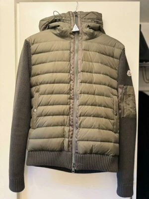 Moncler cardigan - Snygg dunjacka från Moncler i olivgrönt med stickade mörkgrå ärmar och huva. Ej autentisk.Jackan har quiltad front, dragkedja, ribbade muddar och en Moncler-logga på ärmen. Storlek L men passar dig med S då monclers storlekar oftast är mycket mindre.