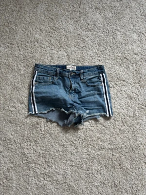 Blå denimshorts med vita ränder - Säljer ett par blå denimshorts från Only med vita dubbla ränder längs sidorna. Shortsens kanter är råa och fransade för en cool look. Klassiska fickor fram och bak samt bälteshällor. Perfekta för sommarens alla äventyr!