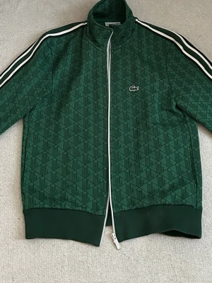 Kofta från Lacoste (fint skick) - Grön zip-tröja från Lacoste med mönstrad design och vita detaljer längs dragkedjan och axlarna.                           Nypris: 1800kr