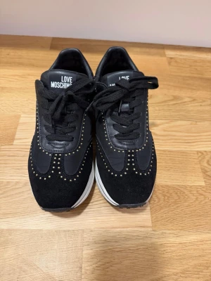 Love Moschino svarta sneakers - Snygga svarta sneakers från Love Moschino med guldfärgade nitar längs kanterna och logga på sidan. Skorna har en mix av mocka och textil, svarta skosnören och vit sula som ger en clean kontrast. Perfekta för dig som vill sticka ut med stil.