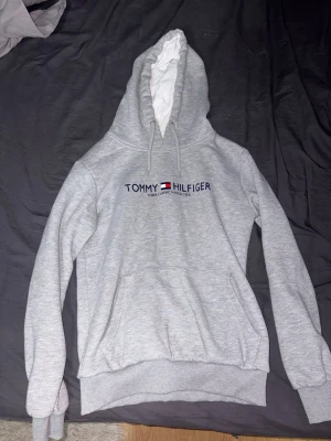 Grå hoodie från Tommy Hilfiger, Storlek M - Snygg ljusgrå hoodie från Tommy Hilfiger med broderad logga och flagga på bröstet. Klassisk känguruficka framtill och justerbar huva med vita snören. Tillverkad i mjukt sweatshirtmaterial, perfekt för en avslappnad stil.