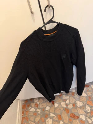 Svart stickad tröja från BOSS - Svart stickad tröja från BOSS med rund halsringning och diskret logotyp på bröstet. Tröjan har en orange detalj på insidan av halsen och är långärmad. Perfekt för dig som gillar stilrena plagg med premiumkänsla.