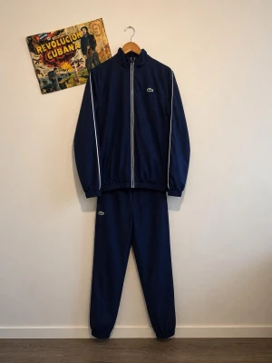 Marinblå tracksuit från Lacoste - Snygg marinblå tracksuit från Lacoste med vita detaljer längs dragkedjan och ärmarna. Jackan har hög krage och stort Lacoste-logotyp i vitt på ryggen. Byxorna har resår i midjan och vid benslut. Perfekt för chill eller streetwear.